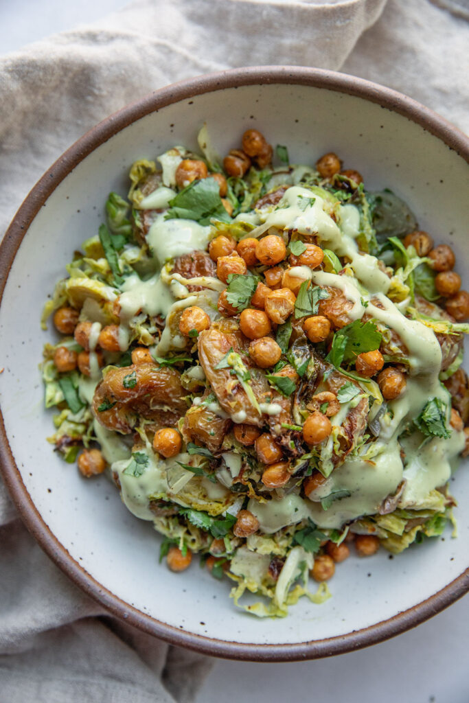 A bowl of roasted brussels sprouts, smashed potatoes and crispy chickpeas topped with avocado dressing.