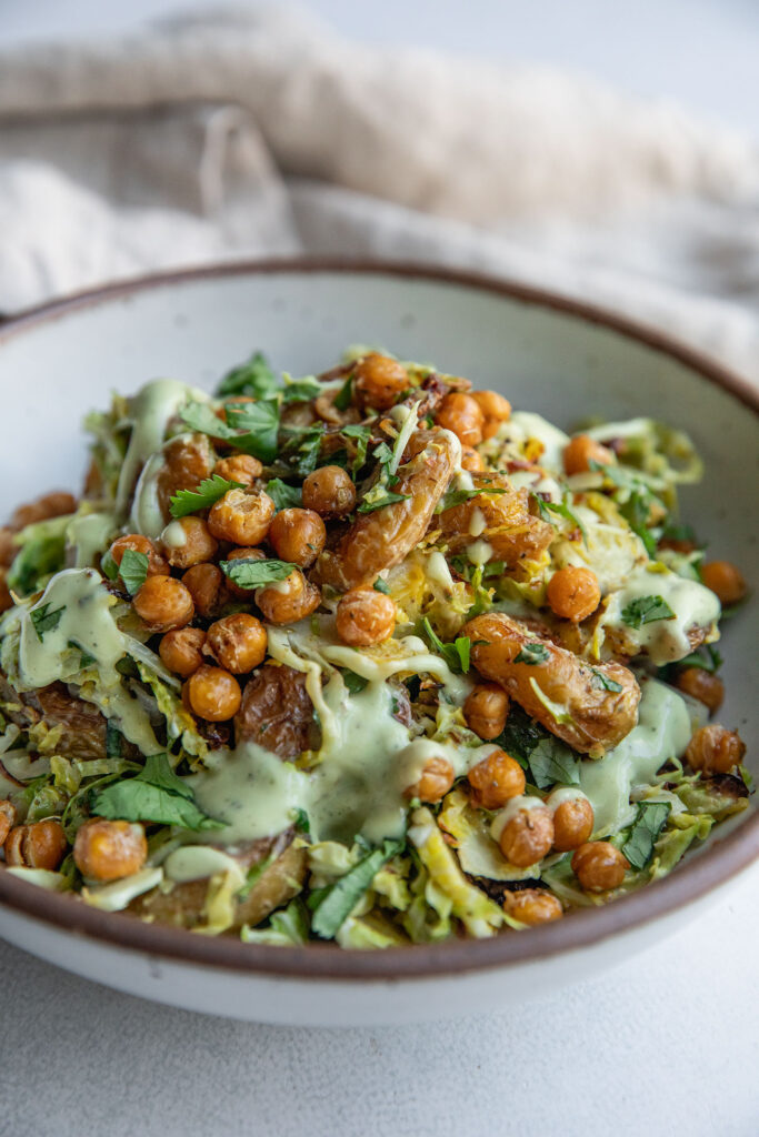 A bowl of brussels sprout salad tossed with roasted potatoes, chickpeas and a creamy avocado dressing.