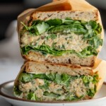 Close up of a halved shishito pepper chickpea salad sandwich layered with lettuce, the chickpea filling, more shishito peppers, cilantro and scallions, served on a white plate.