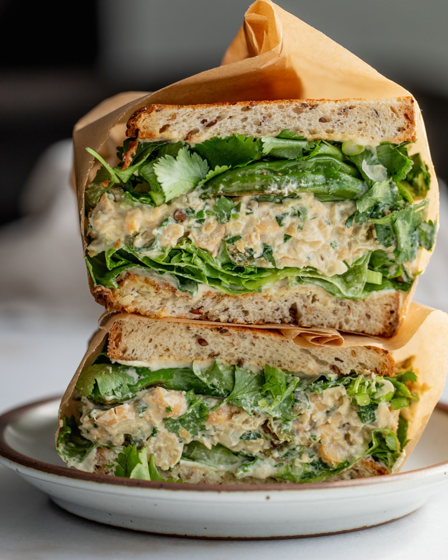 Close up of a halved shishito pepper chickpea salad sandwich layered with lettuce, the chickpea filling, more shishito peppers, cilantro and scallions, served on a white plate.