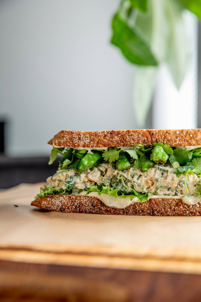 A chickpea salad sandwich on a piece of parchment paper showing a layer of tahini sauce, lettuce, chickpea salad, shishito peppers, cilantro and scallions between two slices of toasted bread.