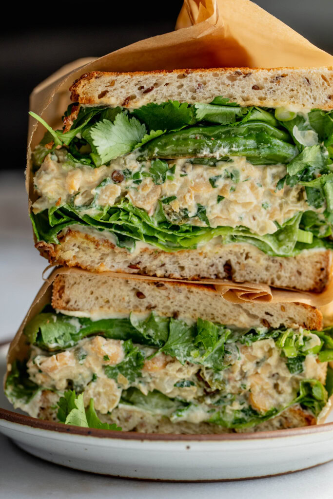 Close up of the sandwich filling showing layers of tahini sauce, lettuce, chickpea salad, shishito peppers, cilantro, and scallions between toasted bread.
