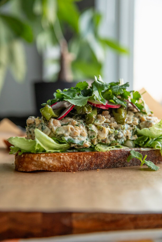 A slice of toasted bread layered with the tahini sauce, lettuce, chickpea salad, shishito peppers, radishes, cilantro, and scallions.