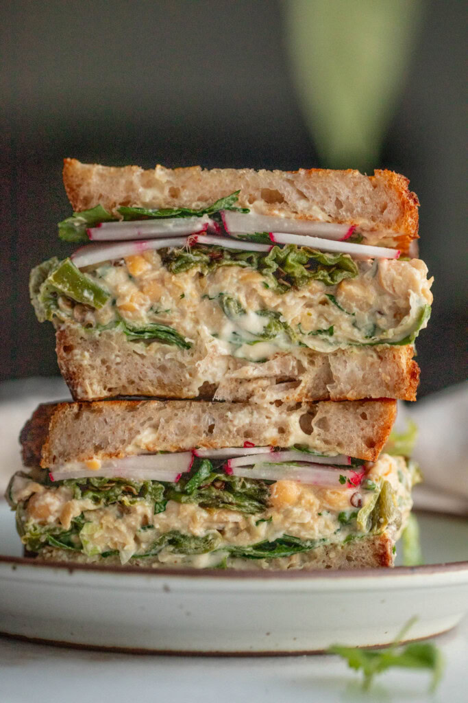 A sandwich cut in half stacked on a plate showing layers of tahini sauce, lettuce, chickpea salad, shishito peppers, radishes, cilantro, and scallions.