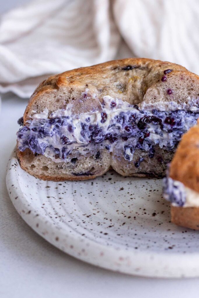 Cross section of a bagel stuffed with cream cheese and blueberry jam on a plate.