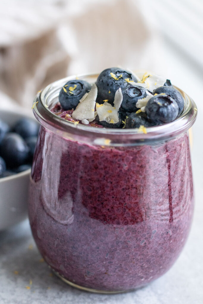 Close up of a jar of chia pudding topped with blueberries, coconut flakes and lemon zest with a bowl of blueberries set behind the jar.