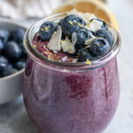 Jar of blueberry chia pudding topped with fresh blueberries, coconut flakes and lemon zest with a bowl of blueberries and lemon set behind the jar.