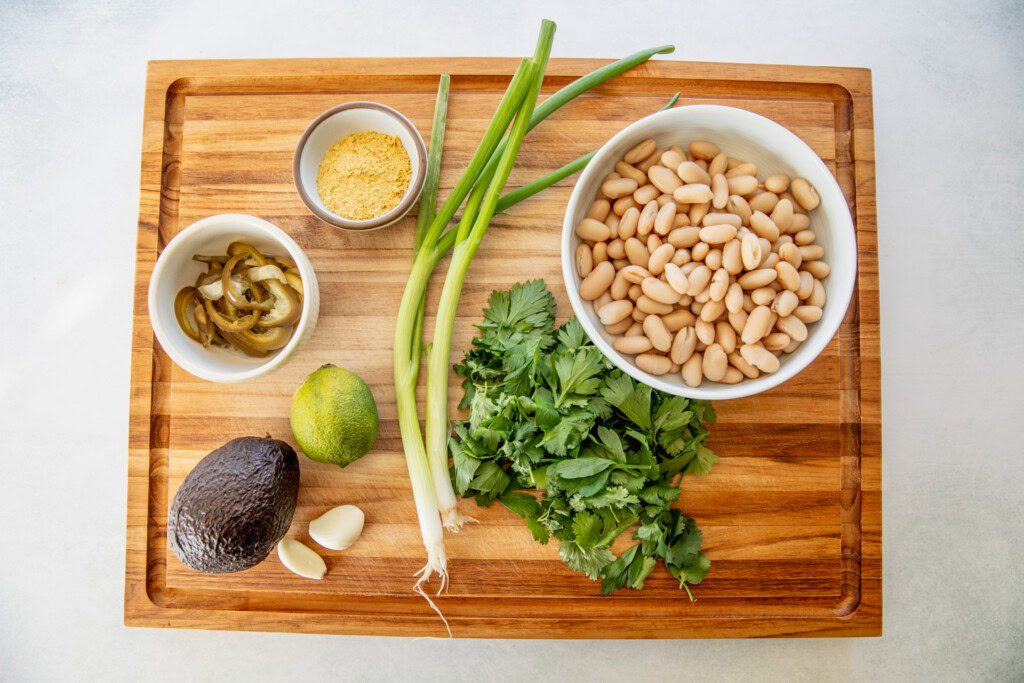 Cutting board topped with bowl of white beans, scallions, garlic cloves, avocado, pickled jalapeños, nutritional yeast, cilantro, parsley, and a lime.