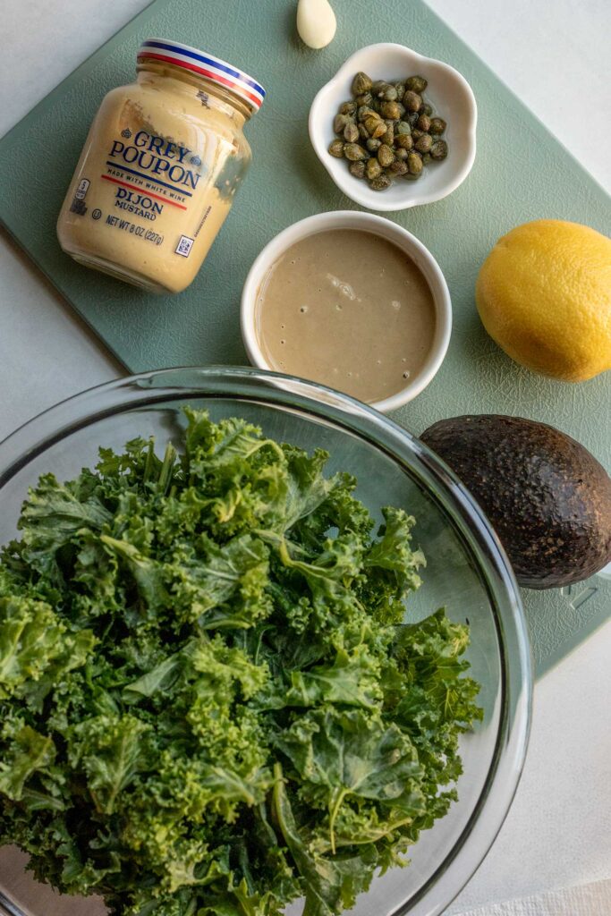 Cutting board with a jar of Dijon mustard, bowl of tahini, avocado, lemon, and capers.