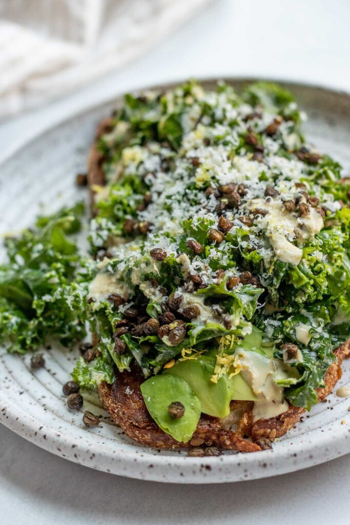 Plate of toast topped with avocado, vegan kale Caesar salad, lentils and grated Brazil nut.