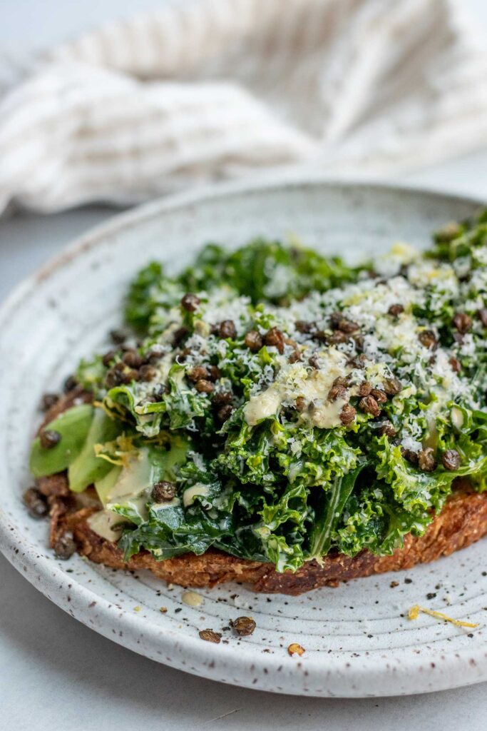 Side view of a plate of toast topped with avocado, kale salad, roasted lentils, Caesar dressing and grated Brazil nut.