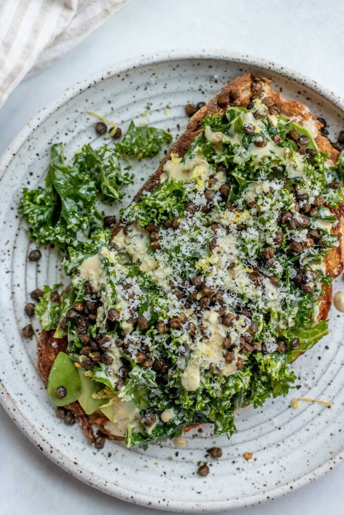 Top down view of avocado toast topped with Caesar salad, roasted lentils and more tahini Caesar dressing.