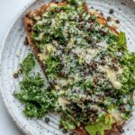 Toast on a white plate topped with avocado slices, Caesar kale salad, roasted lentils and grated Brazil nut.