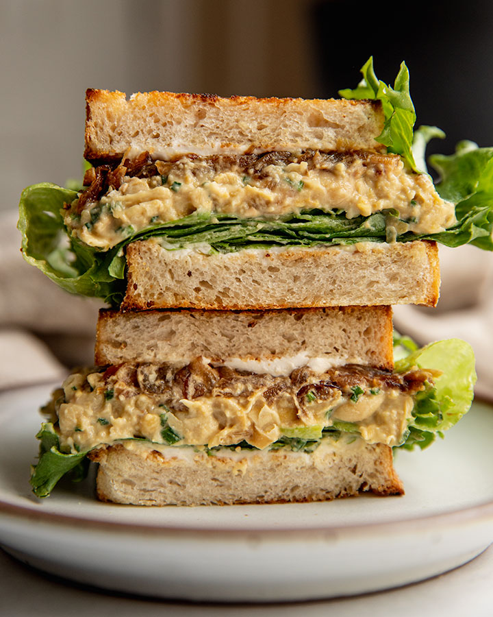 Sandwich cut in half and stacked on top of each other showing layers of lettuce, cream cheese, chickpea salad and caramelized onions.