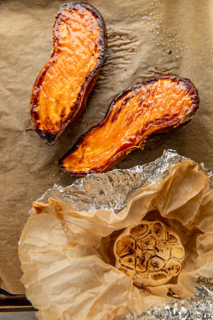 Roasted bulb of garlic and caramelized sweet potato halves on a baking pan.