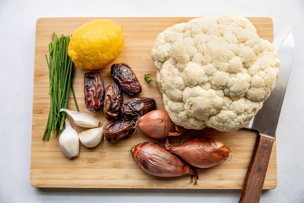 Cutting board topped with shallots, dates, garlic, chives, lemon and a head of cauliflower.