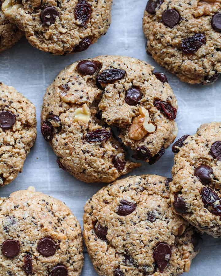 Cookies spread out on parchment paper that are loaded with chocolate chips, raisins and walnuts.