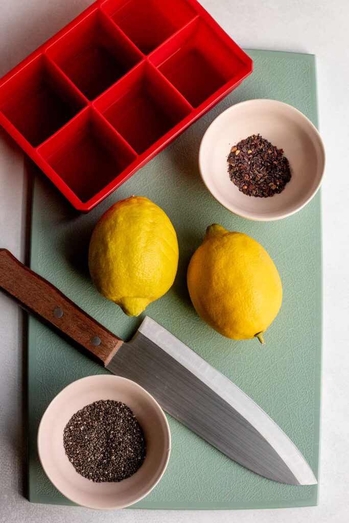 Cutting board topped with ice cube tray, hibiscus leaves, two lemons, and chia seeds.