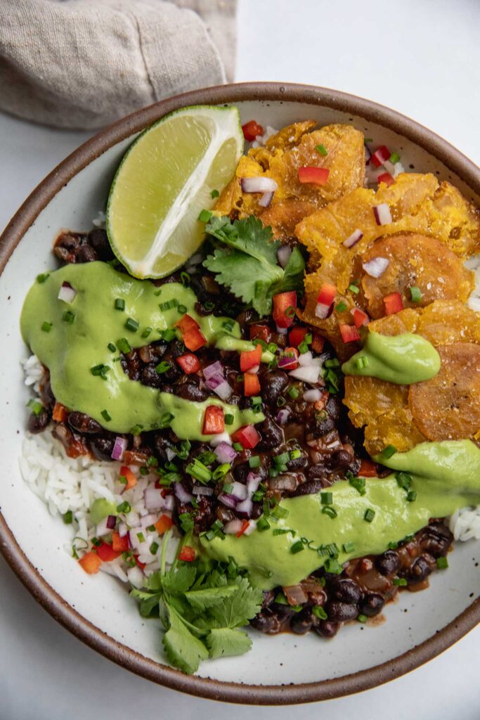 Close up of a bowl of black beans served with rice and topped with avocado cilantro sauce and a side of plantains.