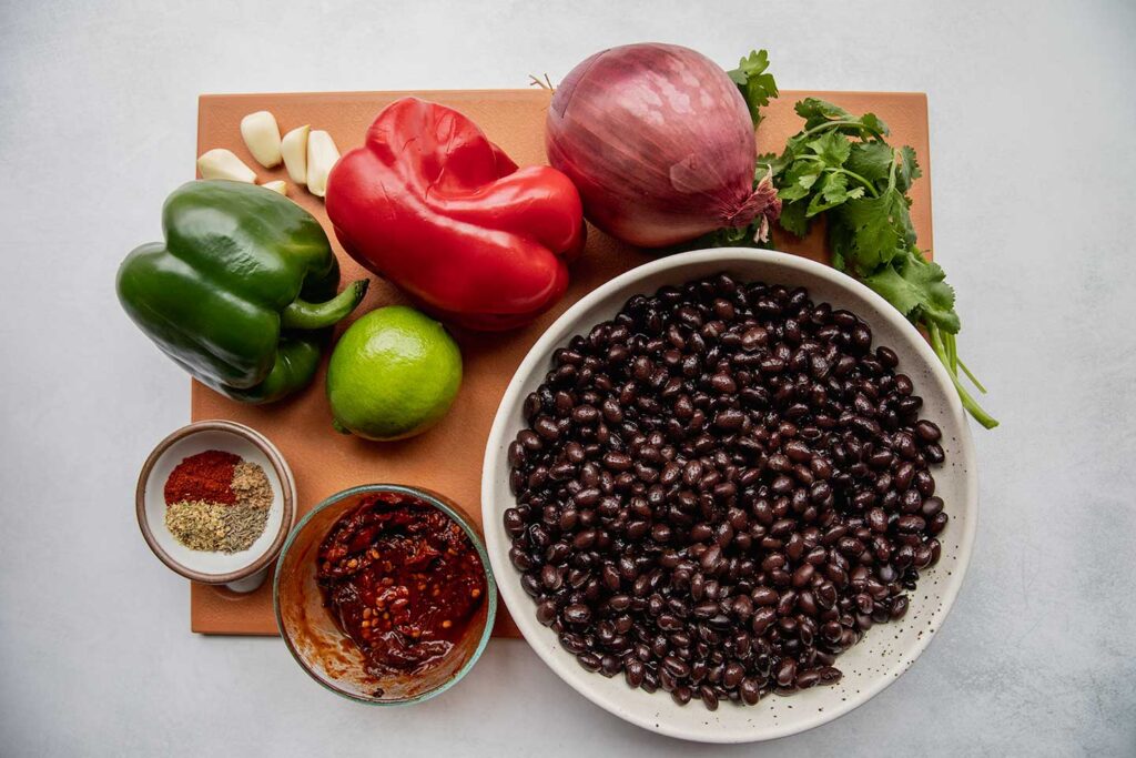 Cutting board topped with green and red bell pepper, garlic, lime, chipotle peppers, spices, cilantro, and a bowl of black beans.