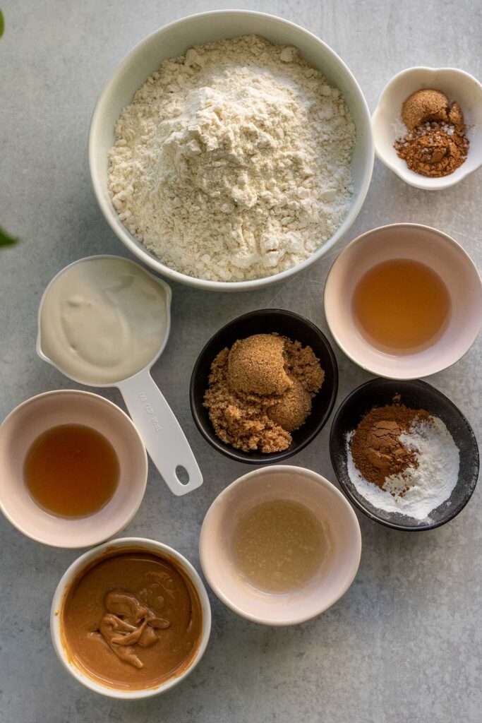 Flour, brown sugar, cinnamon, apple cider vinegar, yogurt, maple syrup, almond butter in separate bowls on a working surface.