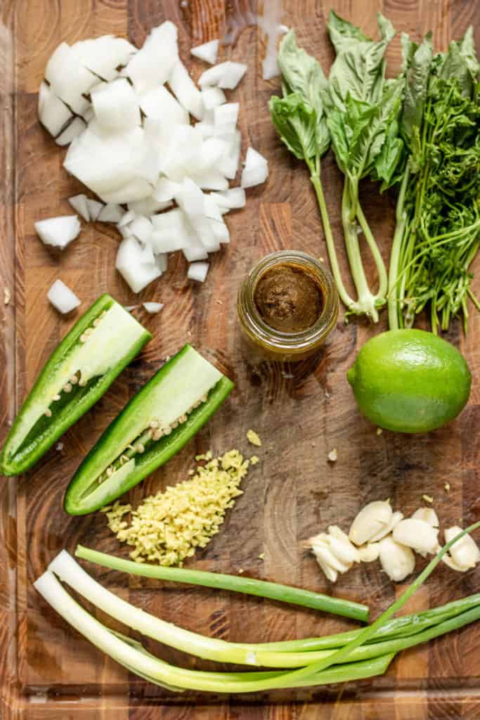 Cutting board with some diced onion, fresh herbs, lime, curry paste, jalapeno, ginger, garlic and scallions.