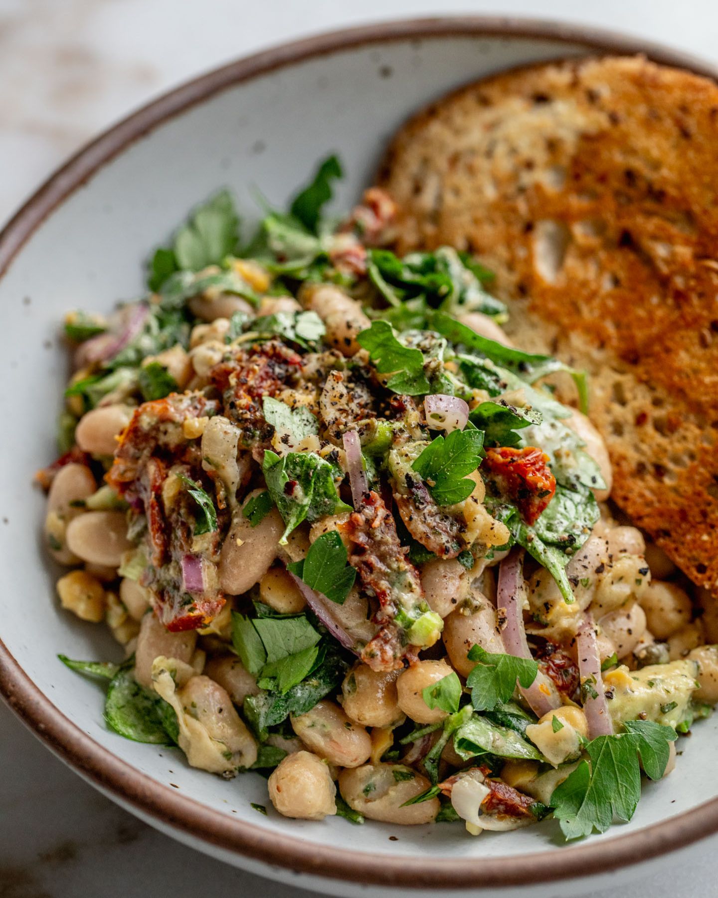 Close up of a bowl of bean salad with a piece of toasted bread in the background.