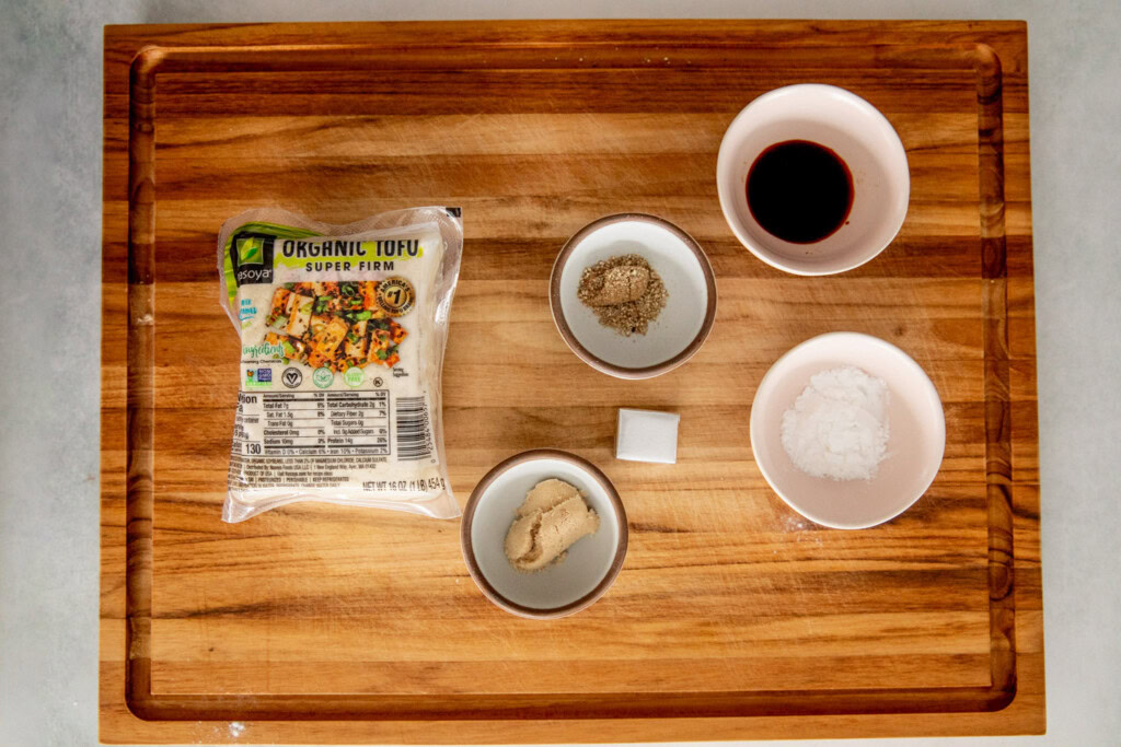 Cutting board topped with super firm tofu, bowl of seasonings, brown sugar, bouillon cube, potato starch and soy sauce.