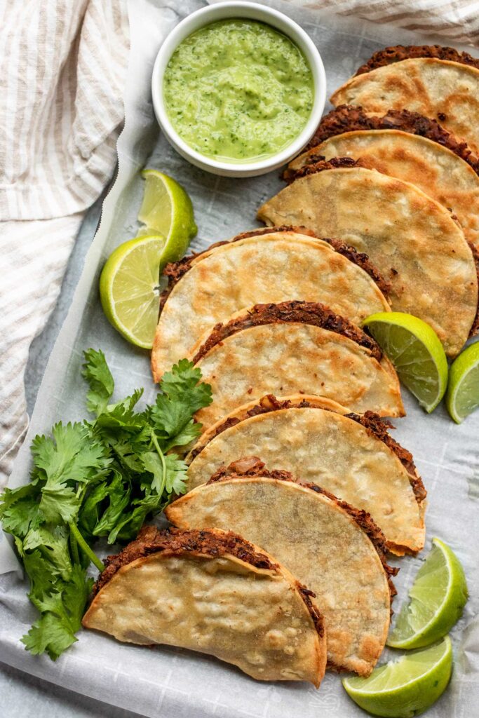A side view of a row of crispy black bean tacos served with cilantro lime sauce arranged on a baking tray with lime wedges and cilantro.