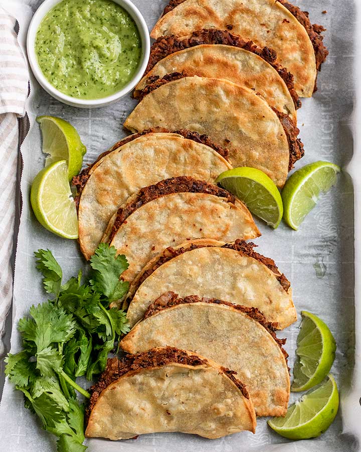 Chipotle black bean stuffed tacos arranged on a baking tray with lime wedges and cilantro.