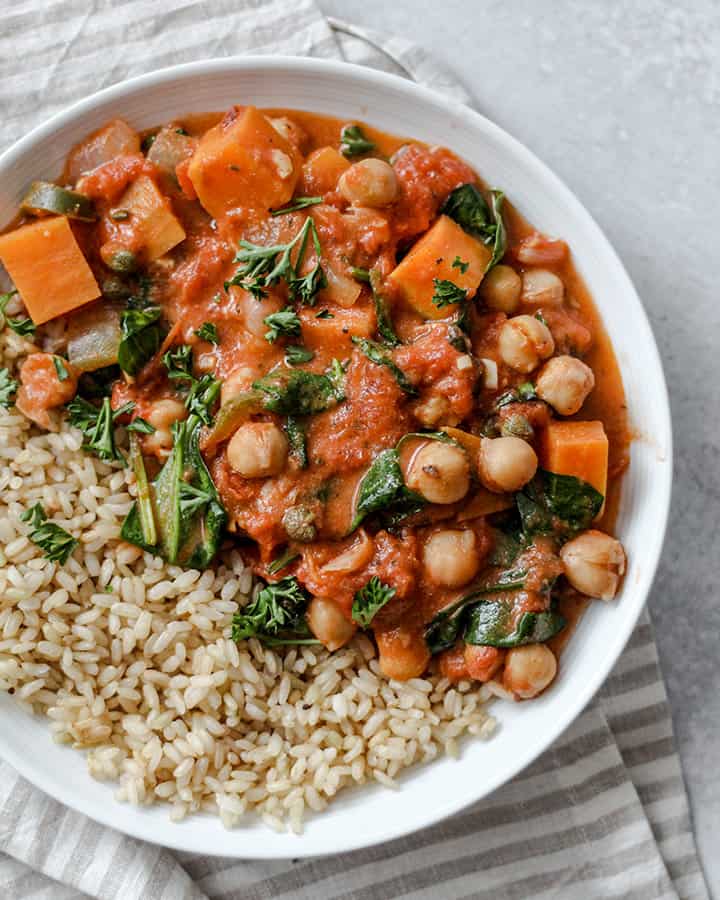 Brown rice paired with the tomato stew and topped with parsley against a grey background and stripped napkin behind.