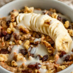 Bowl of cooked oats topped with banana, raisins, walnuts, and cream cheese frosting.