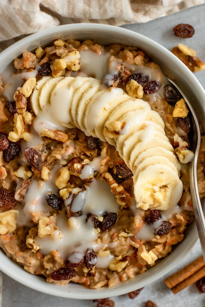 Close up of a bowl of carrot cake oats topped with banana slices, raisins, walnuts and cream cheese frosting with a spoon tucked on the side.
