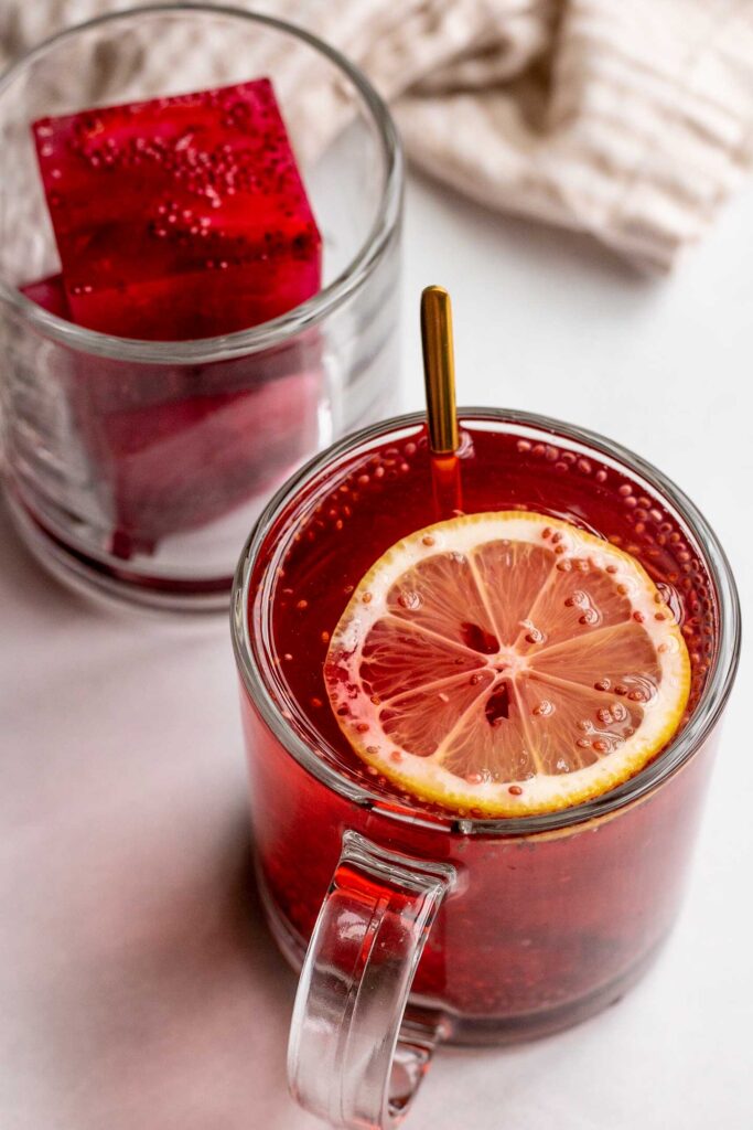 One mug in the background with two hibiscus ice cubes stacked on top of each other and the front mug with boiling water poured over the ice cubes with a lemon slice floating on top.