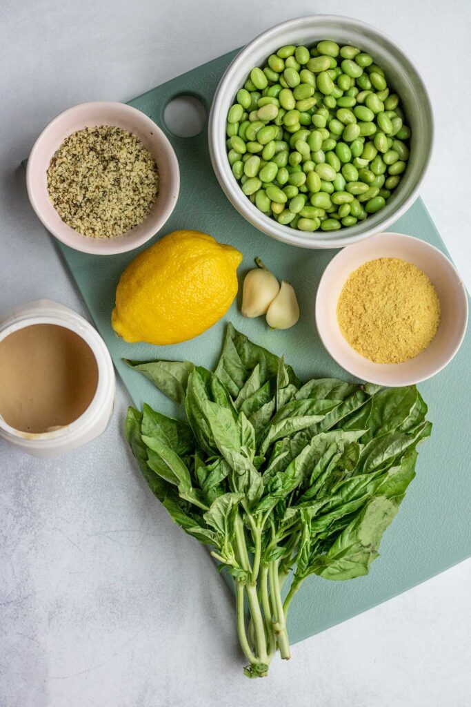 Cutting board topped with basil, nutritional yeast, hemp hearts, edamame, lemon, garlic cloves and tahini.