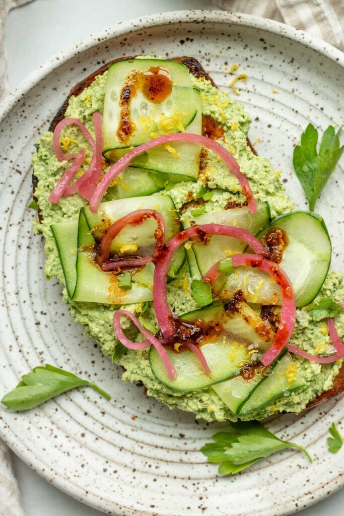 Close up of a high protein toast spread with an edamame spread and topped with cucumber slices, pickled red onion, lemon zest and chili oil.