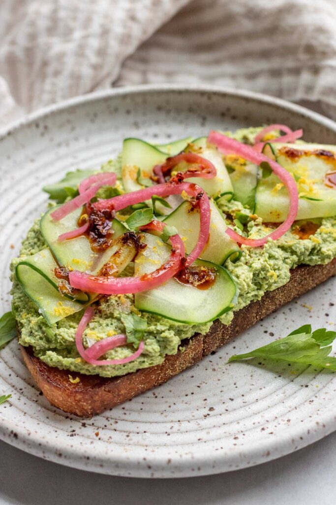 Plate of toast spread with edamame mash, cucumber slices, pickled onion, chili oil and lemon zest.