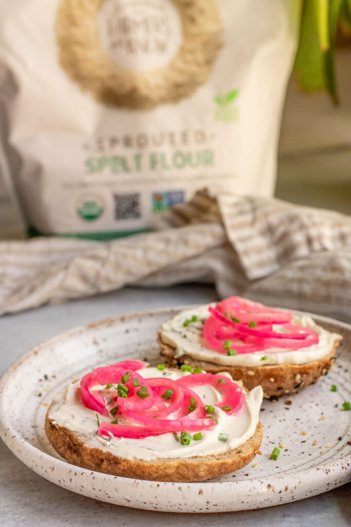 Bag of spelt flour in the background of a plate with a bagel spread with vegan cream cheese topped with pickled onions and chives.