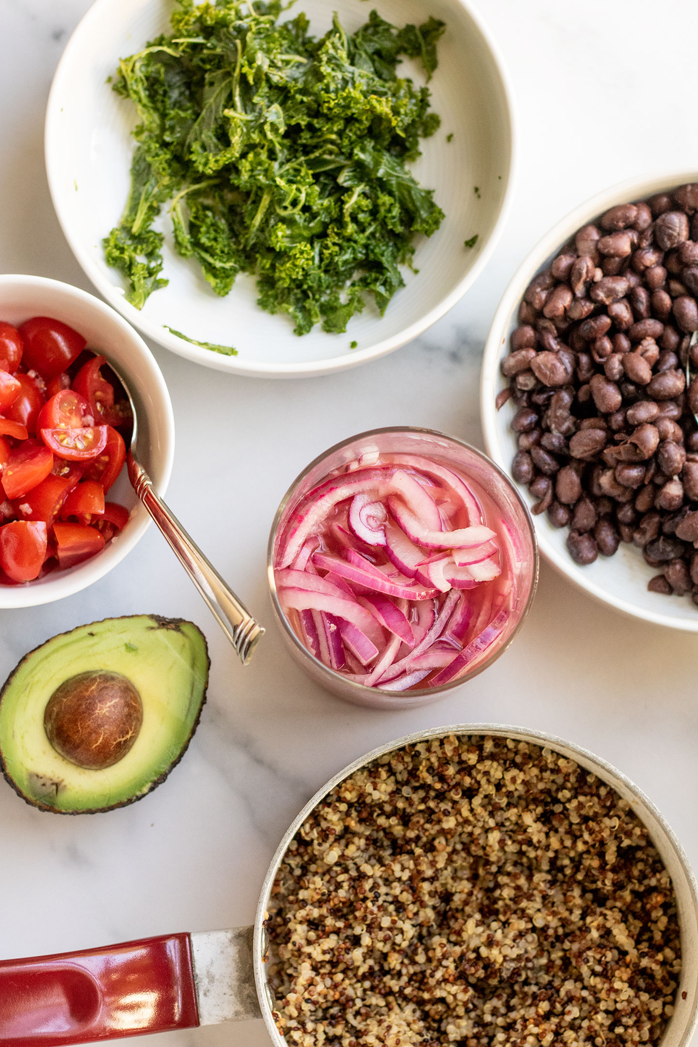 Counter filled with bowls of breakfast jar assembly ingredients including cherry tomatoes, massaged kale, black beans, pickled onions, avocado and quinoa in a pot.