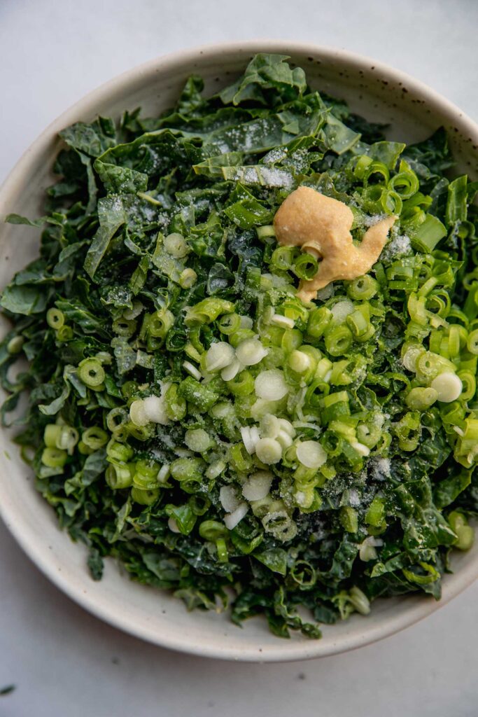 Kale, scallions, mustard, maple syrup and oil in a white bowl.