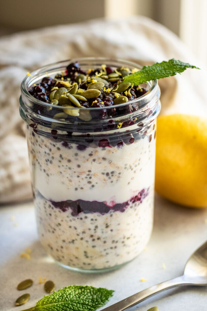 Jar of oats placed on a white flat surface, topped with blueberry jam, pepitas and lemon zest.