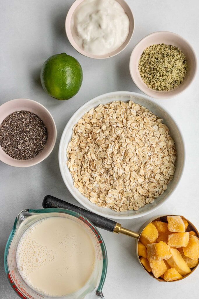 Bowl of rolled oats, frozen mango, lime, soy milk, and chia seeds on a working surface.
