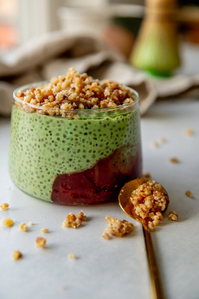 Matcha pudding in a jar with a dollop of strawberry jam topped with pie crust crumble and a gold spoon set next to it.