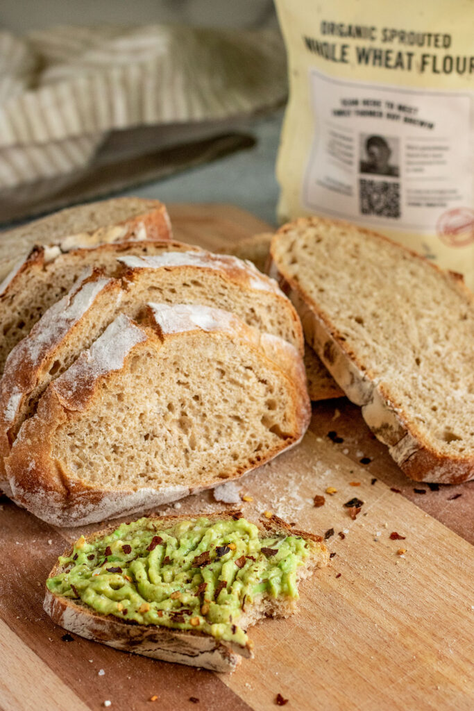 Whole wheat bread slices on a cutting board with one slice spread with mashed avocado and a bag of whole wheat flour in the background.