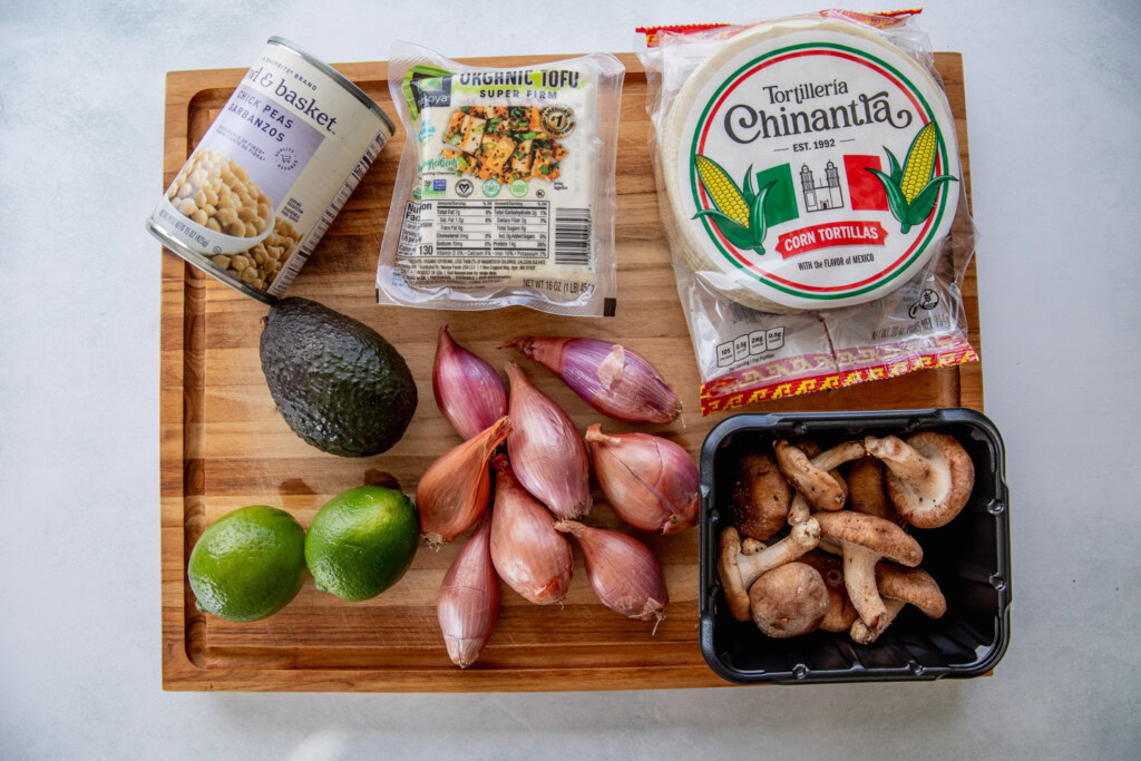 Cutting board with tortillas, super firm tofu, can of chickpeas, avocado, 2 limes, shallots, and a container of shiitake mushrooms.