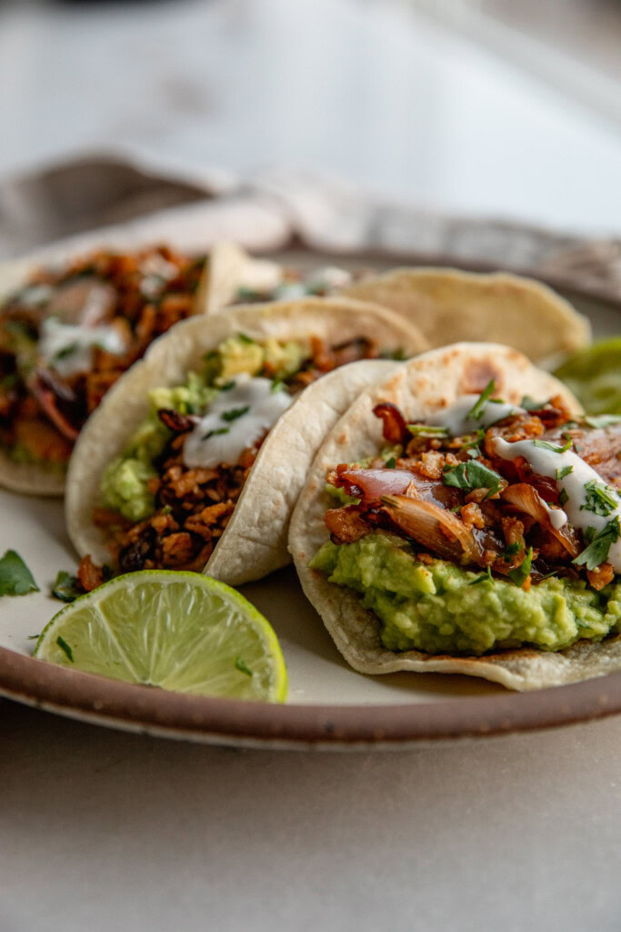 Plate of 4 tortillas stuffed with avocado and shredded tofu and topped with crema served with a 2 wedges of lime.