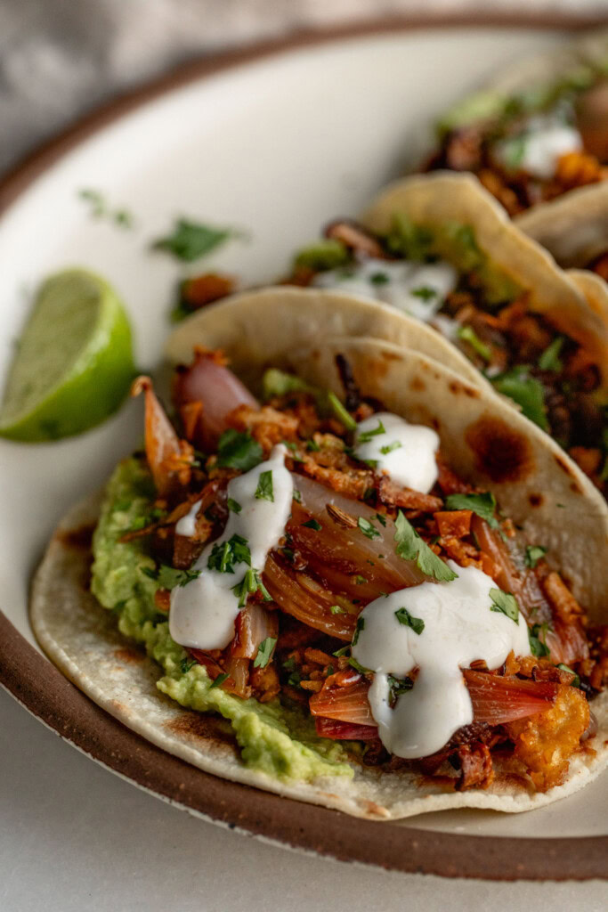 Close up of a taco filled with mashed avocado, shredded tofu and onions with a drizzle of crema on top with a wedge of lime in the background.