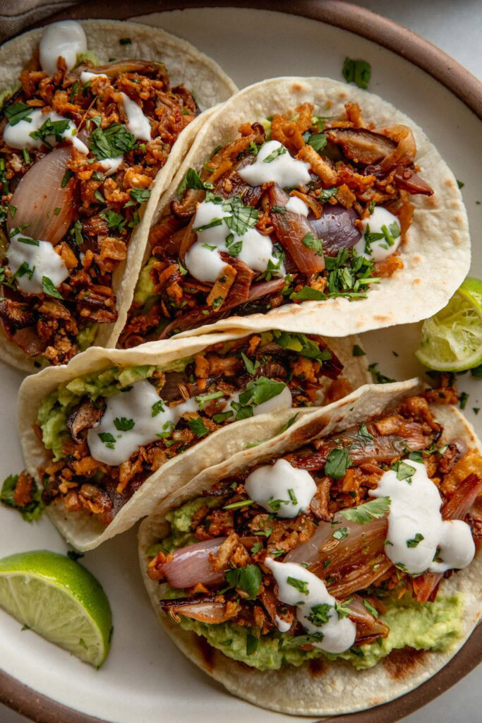 Four tacos lined on a plate stuffed with mashed avocado and shredded tofu and crema served with 2 wedges of lime on the side.