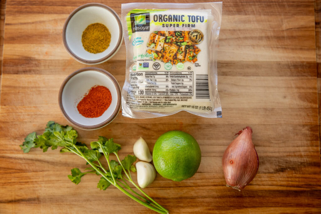 Cutting board topped with a block of tofu, two bowls of spices, garlic, lime, shallot, and a sprig of cilantro.