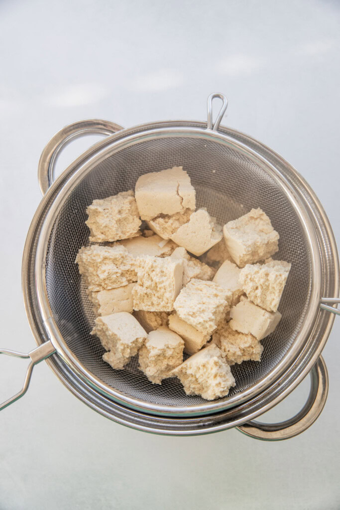 Tofu draining in a metal sieve over a pot.